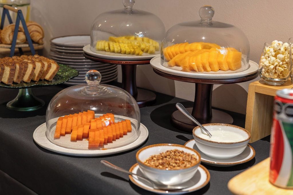 A buffet with various food items including a covered dish of sliced fruit, a bowl of cereal, and a container of popcorn.