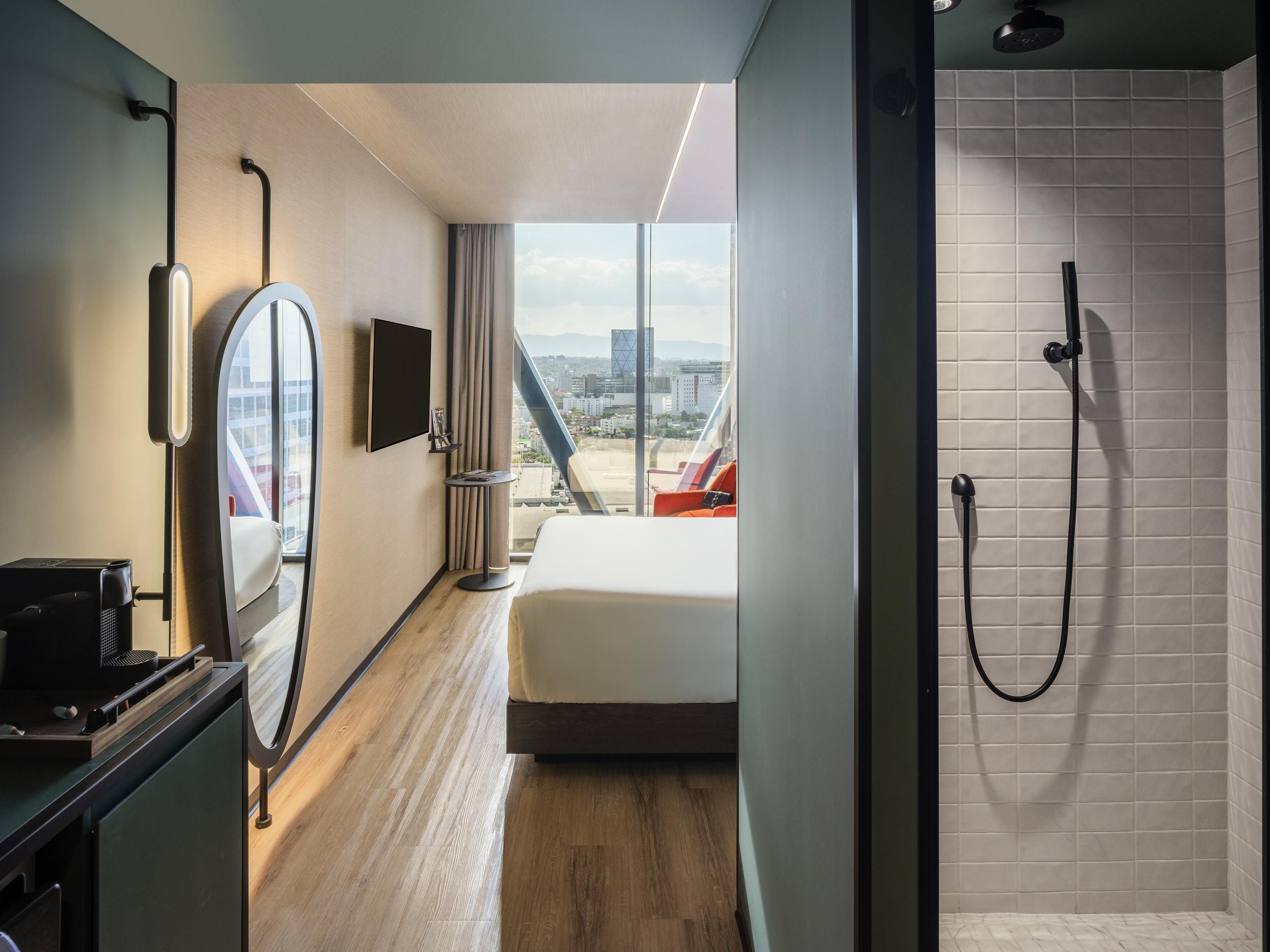 A modern hotel room with a bed, a TV, a desk with a coffee maker, a shower, and a city view.