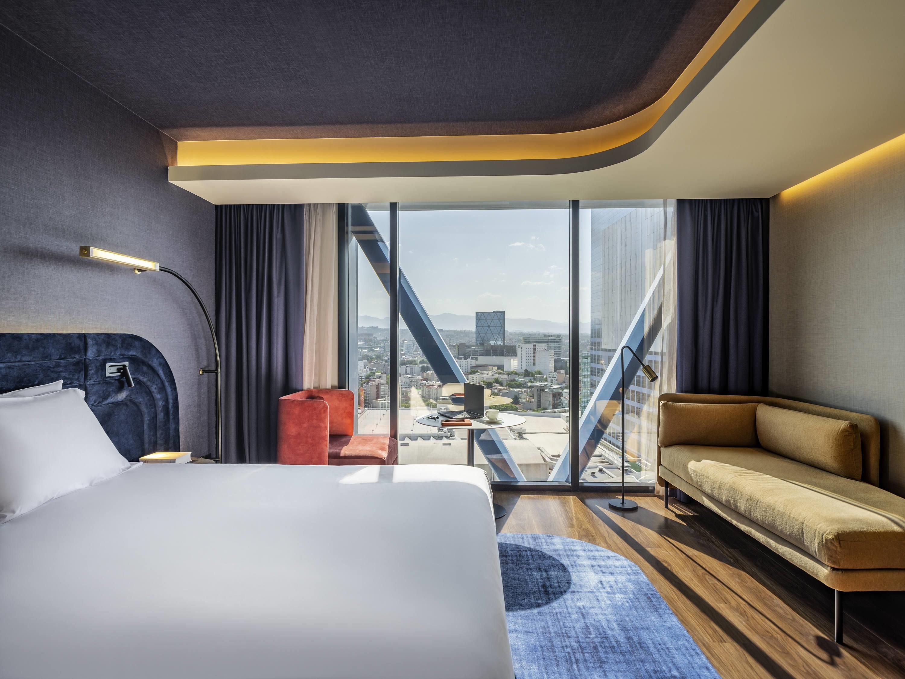 Ett modernt hotellrum med en stor säng, en soffa, ett litet bord och utsikt över stadsbilden genom stora fönster.