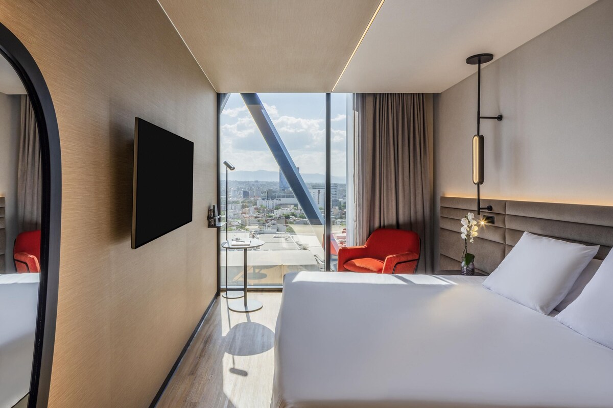 Hotelkamer met een bed, een aan de muur bevestigde televisie, een bureau met stoel, en uitzicht op de stad door grote ramen.