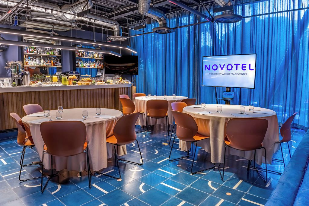 Un bar moderno de hotel con mesas y sillas redondas, una barra con varias botellas y una pantalla que muestra 'NOVOTEL'.