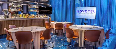 Un bar moderno de hotel con mesas y sillas redondas, una barra con varias botellas y una pantalla que muestra 'NOVOTEL'.