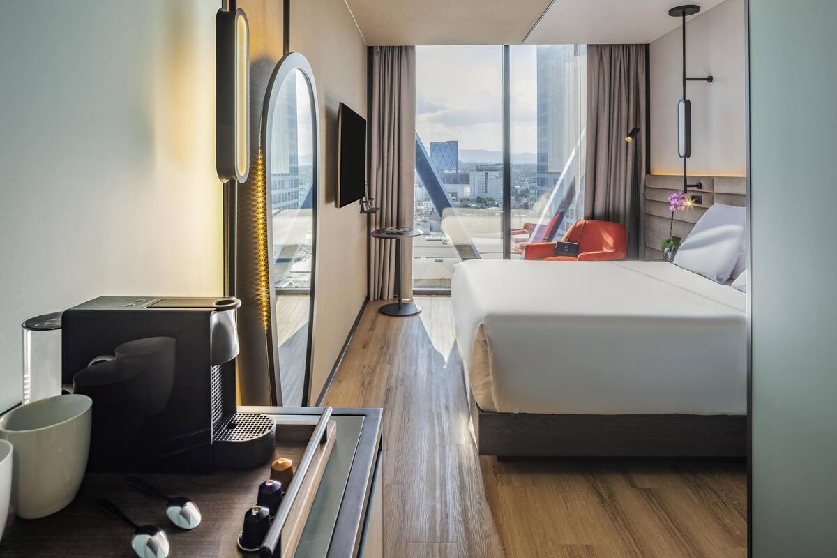 Habitación de hotel con cama, escritorio con cafetera, televisión y vista a la ciudad.