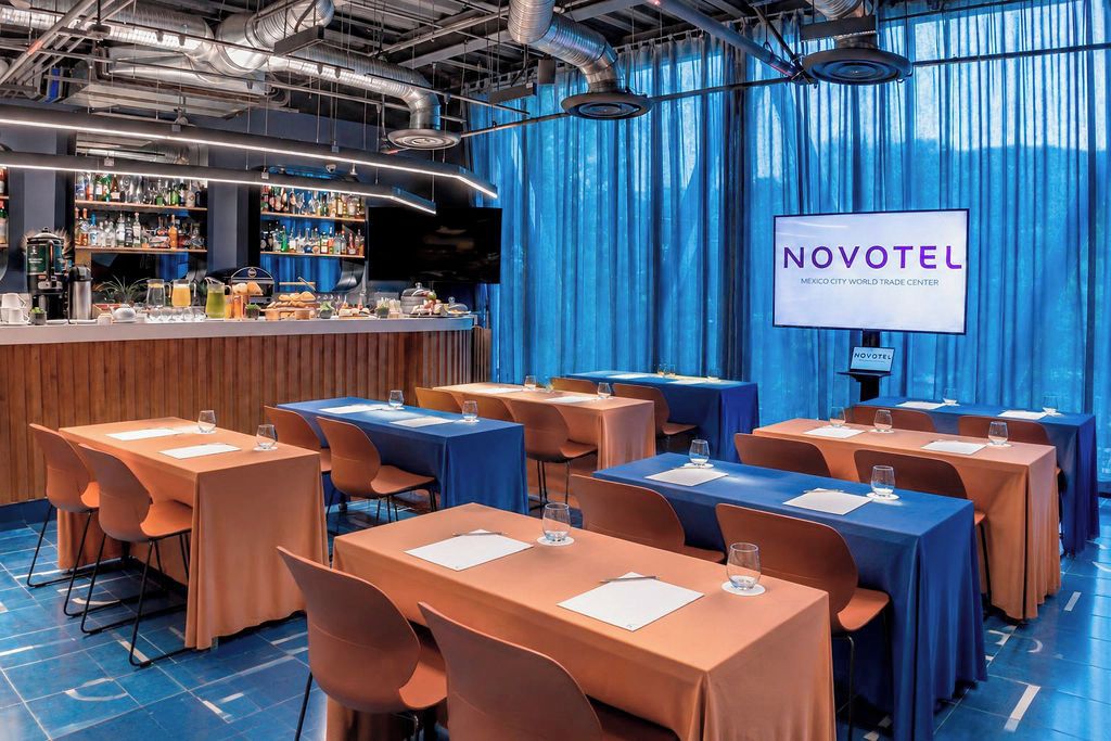 A hotel conference room with tables, chairs, a bar area, and a screen displaying the hotel name.