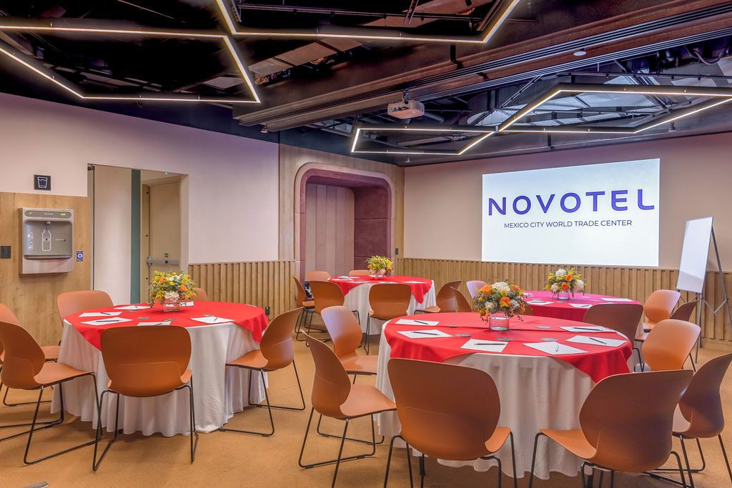 A conference room with multiple tables and chairs, a projector screen, and a sign reading 'NOVOTEL MEXICO CITY WORLD TRADE CENTER'.