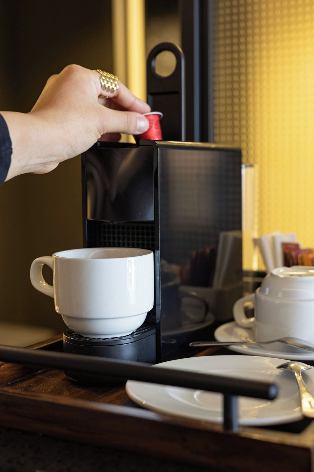 Eine Hand legt eine weiße Tasse auf ein Tablett und in eine schwarze Kaffeemaschine.