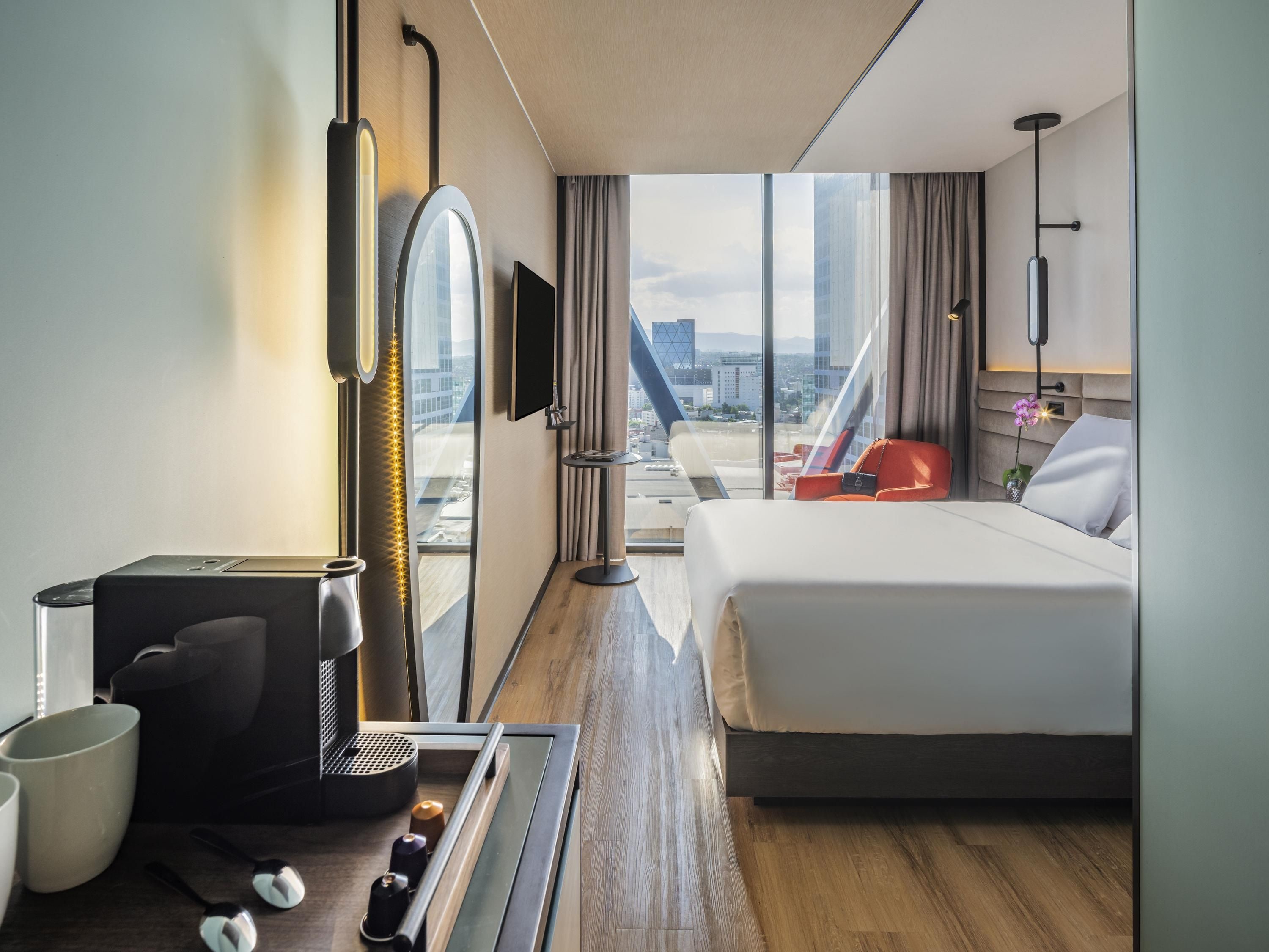 A hotel room with a bed, a desk with a coffee machine, a TV, and a view of the city.