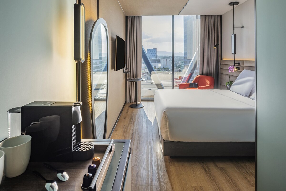 Hotelkamer met een bed, een bureau met een koffieapparaat, een televisie en uitzicht op de stad.
