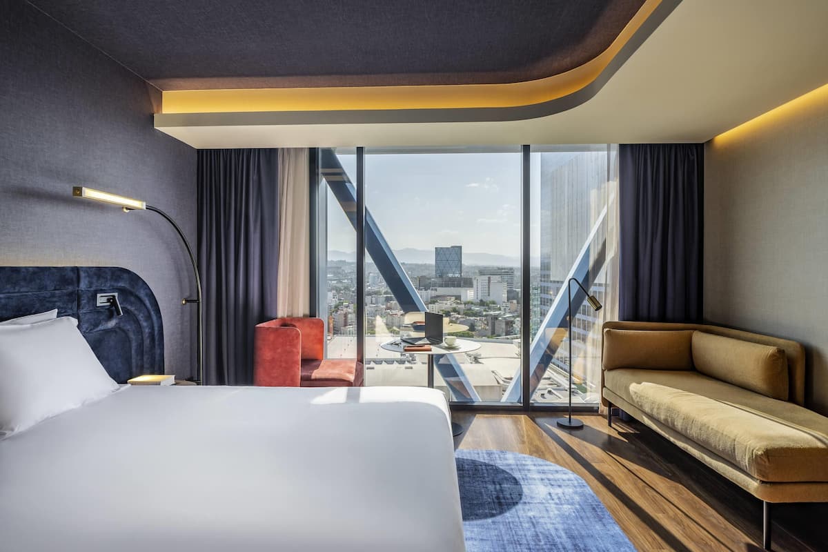 Habitación de hotel moderna con una cama grande, un sofá, una mesita y vistas de la ciudad a través de amplias ventanas.