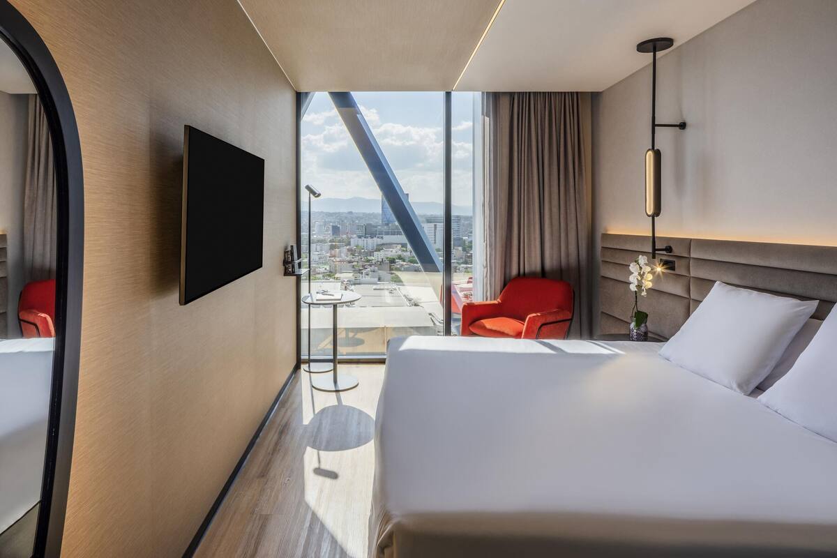 Habitación de hotel con una cama, un televisor en la pared, un escritorio con una silla y vistas a la ciudad a través de amplias ventanas.