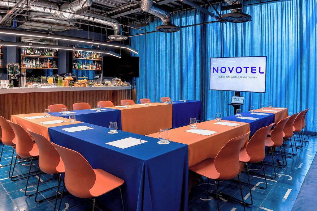 Un salón de conferencias con mesas y sillas, una zona de bar con varias botellas y una pantalla que muestra 'NOVOTEL'.
