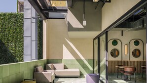 A modern outdoor seating area with cushioned benches and a table, surrounded by a glass wall with decorative mirrors and a green hedge. - Novotel Mexico City World Trade Center (Mexico City)