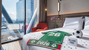 A hotel room with a bed, a red chair, a glass table, a view of a cityscape, and a welcome sign. - Novotel Mexico City World Trade Center (Mexico City)