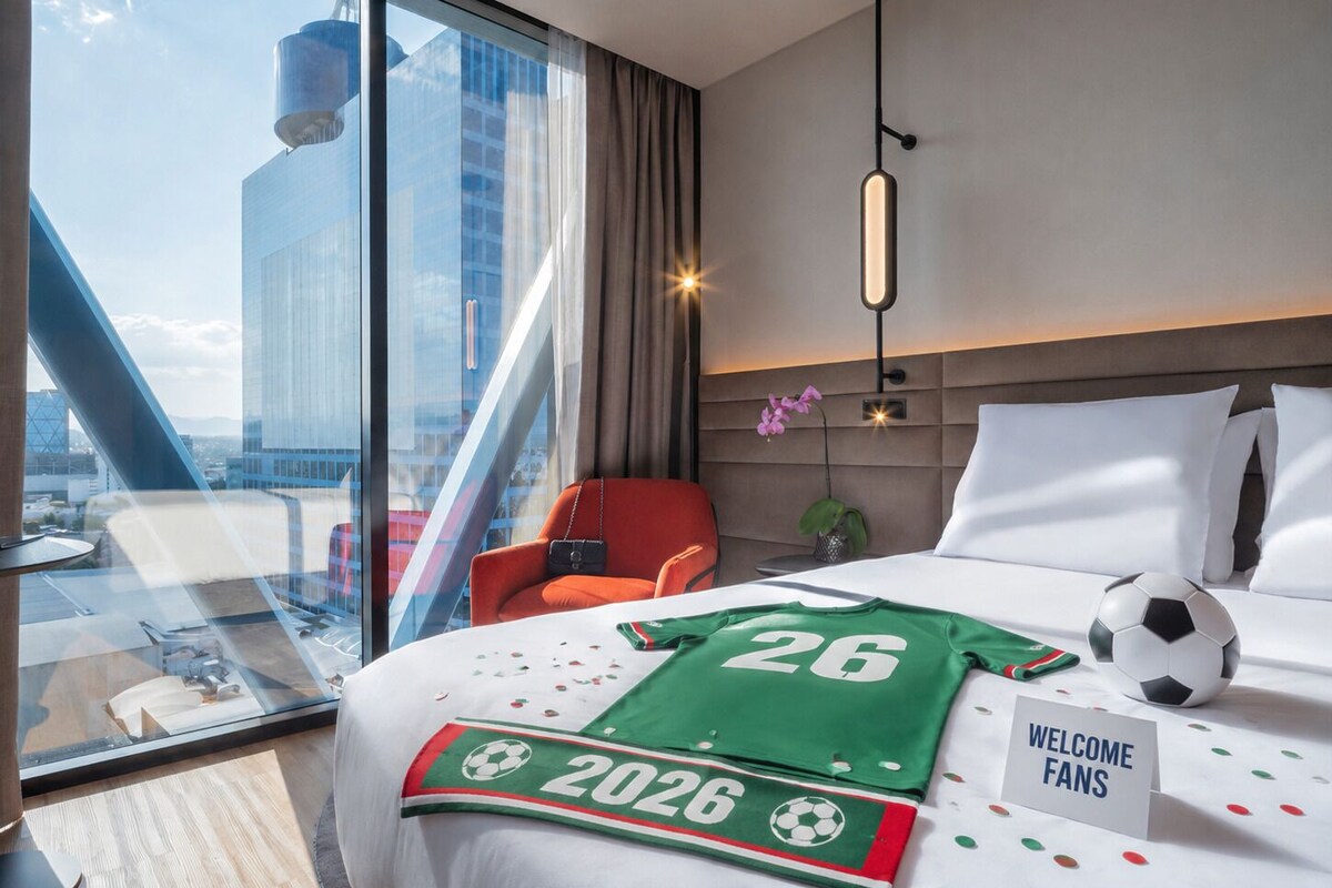 A hotel room with a bed, a red chair, a glass table, a view of a cityscape, and a welcome sign.