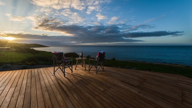 Una terraza de madera con dos sillas y una mesa, con vistas a un cuerpo de agua y un paisaje cubierto de césped al atardecer.