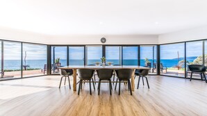 En rummelig spiseplads med et træbord og stole, store vinduer med havudsigt og et moderne ur på væggen.