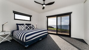 Et soveværelse med seng, natbord, skrivebordslampe, loftventilator og en stor skydedør, der fører ud til en balkon med havudsigt.