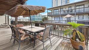 Um deck de madeira com mesa e cadeiras, um guarda-sol de palha e um vaso com planta.