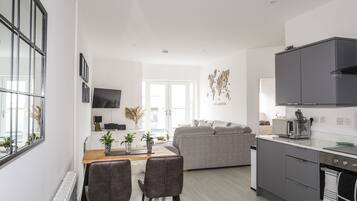 A modern kitchen with grey cabinets, a built-in oven, and a microwave. There is a dining area with a wooden table and chairs, and a grey sofa in the background.