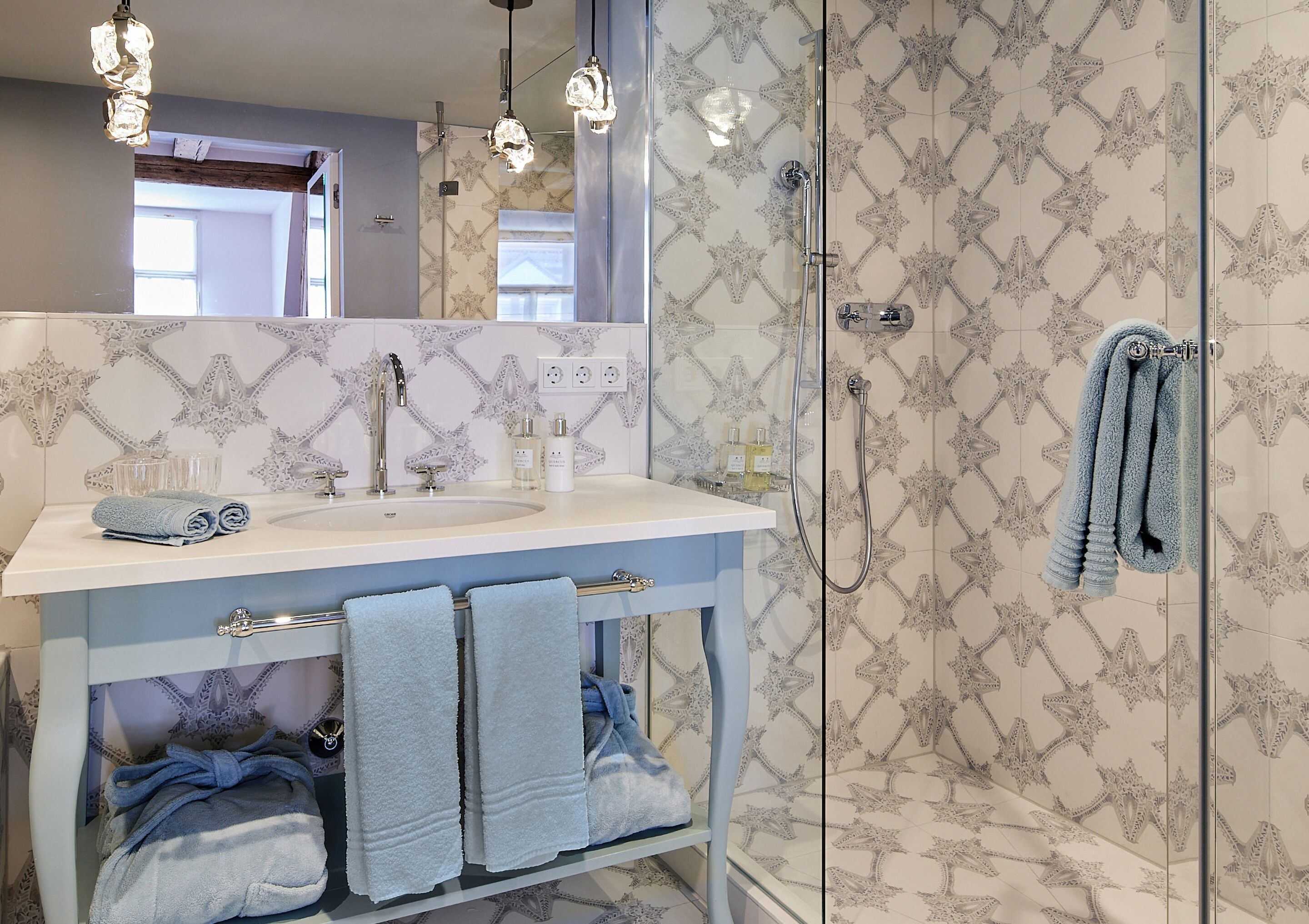 A bathroom with a white sink, two towels, a glass shower enclosure, and patterned wall tiles.