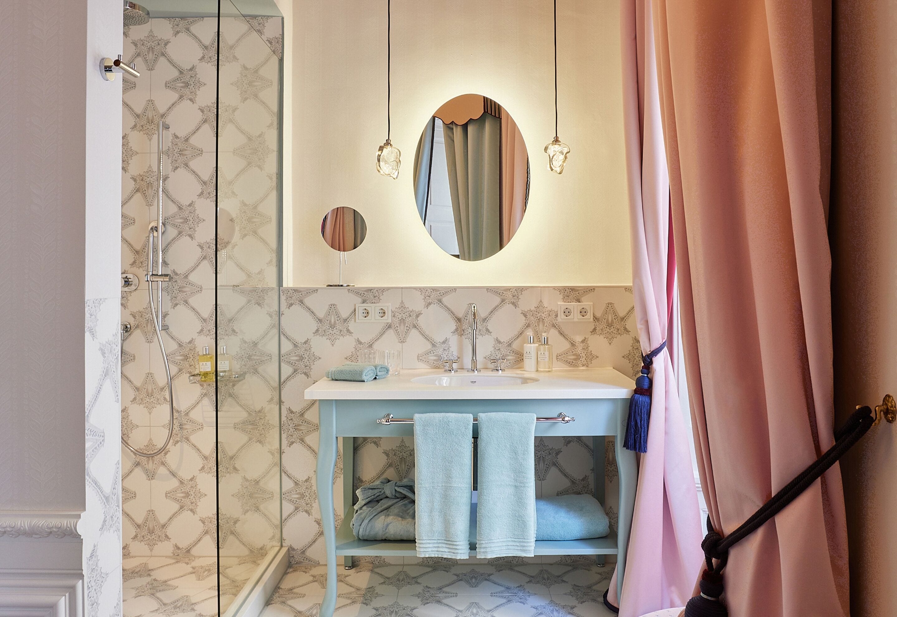 Un baño con ducha, lavabo y espejo. Hay toallas colgadas en un perchero y una cortina parcialmente abierta.