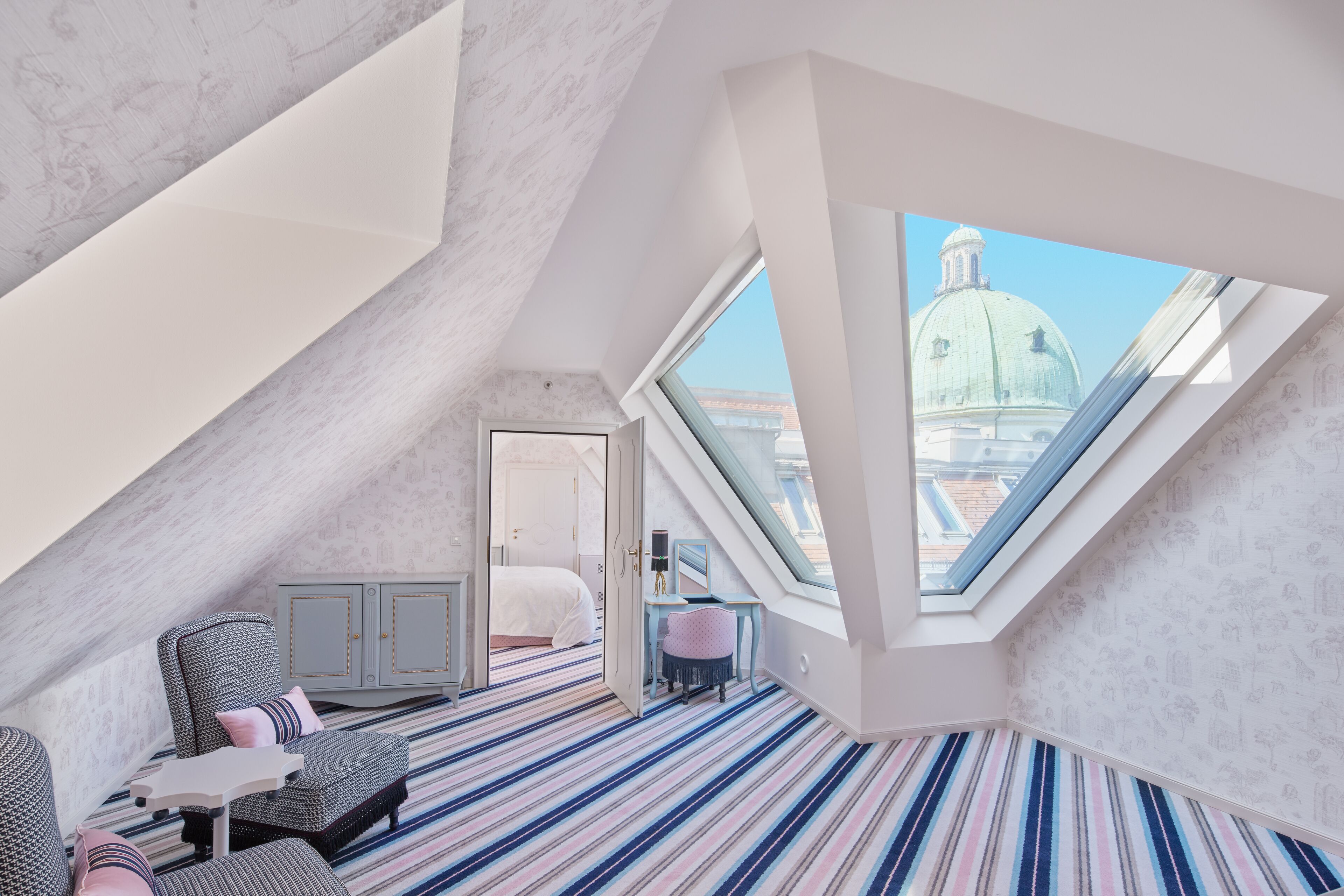 A modern loft bedroom with a sloped ceiling, a large skylight, a striped carpet, a bed, a desk, and a view of a dome building.