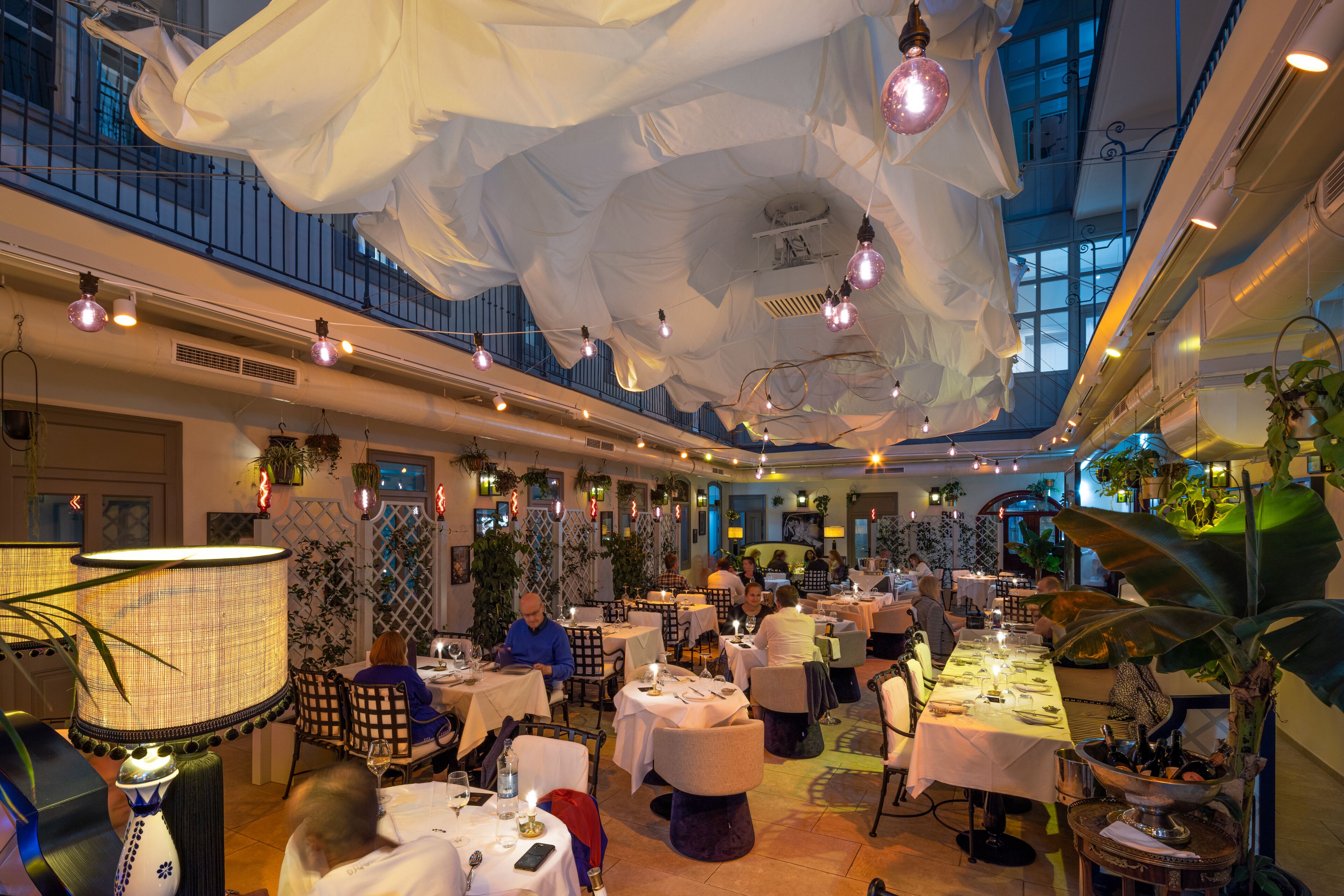 Un restaurant avec des tables dressées pour le dîner, un plafond haut orné de luminaires suspendus et une vue sur le ciel grâce à de grandes fenêtres.
