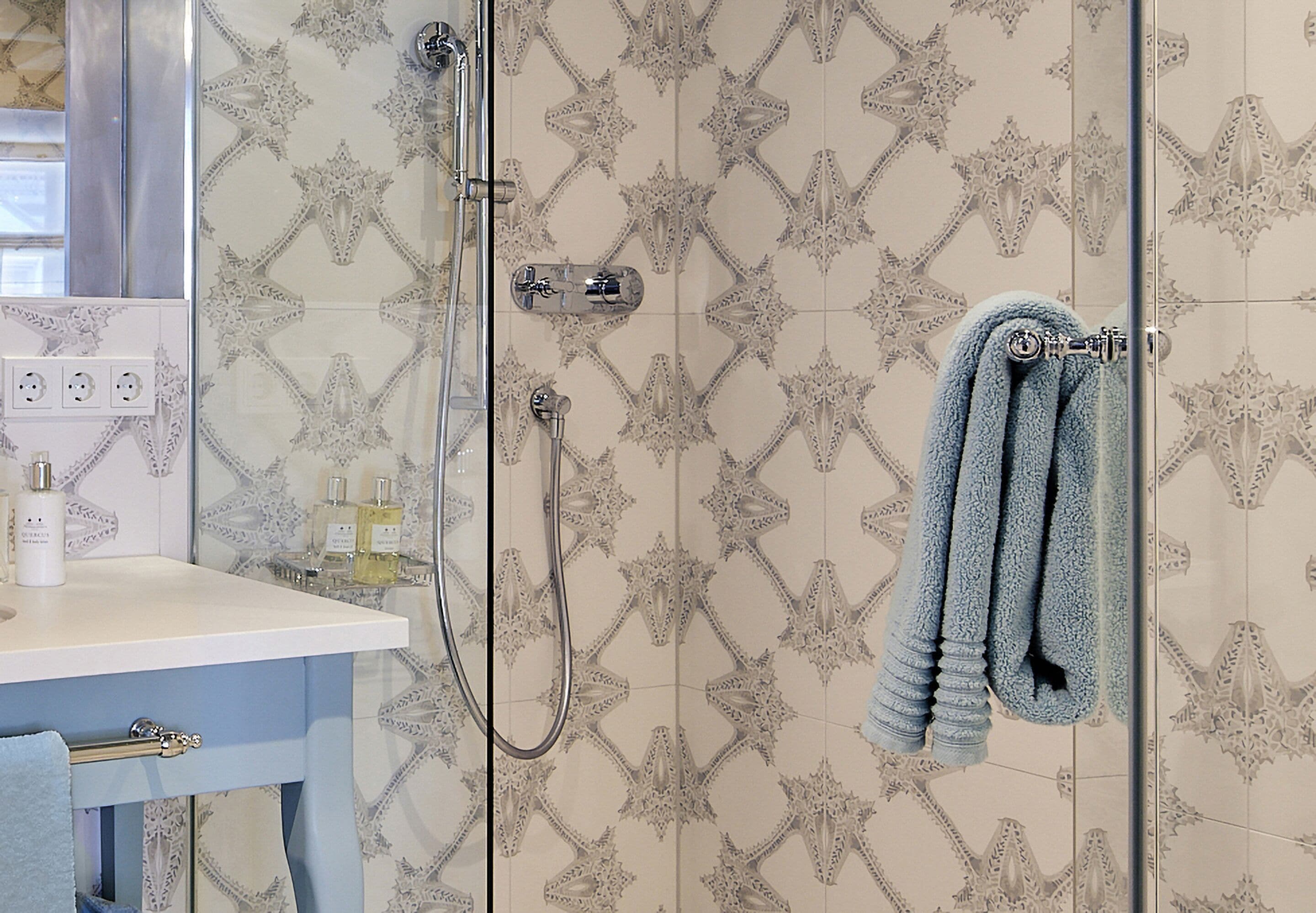 Ein Badezimmer mit gemusterten Fliesen, einer Duschkabine aus Glas, einem Handtuchhalter mit Handtüchern, einem Waschbecken mit Toilettenartikeln und einem Spiegel.