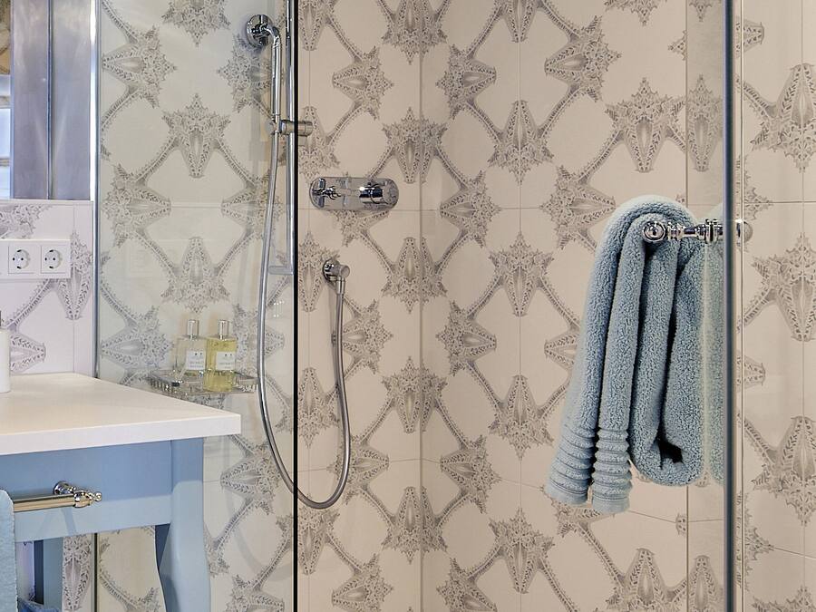 Ein Badezimmer mit gemusterten Fliesen, einer Duschkabine aus Glas, einem Handtuchhalter mit Handtüchern, einem Waschbecken mit Toilettenartikeln und einem Spiegel.