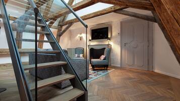 A modern loft bedroom with exposed wooden beams, a glass staircase, and a cozy seating area with a fireplace.