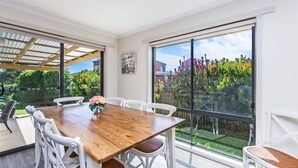 Et spiseområde med et træbord og hvide stole, store vinduer med udsigt til en have og et loft med ovenlys.