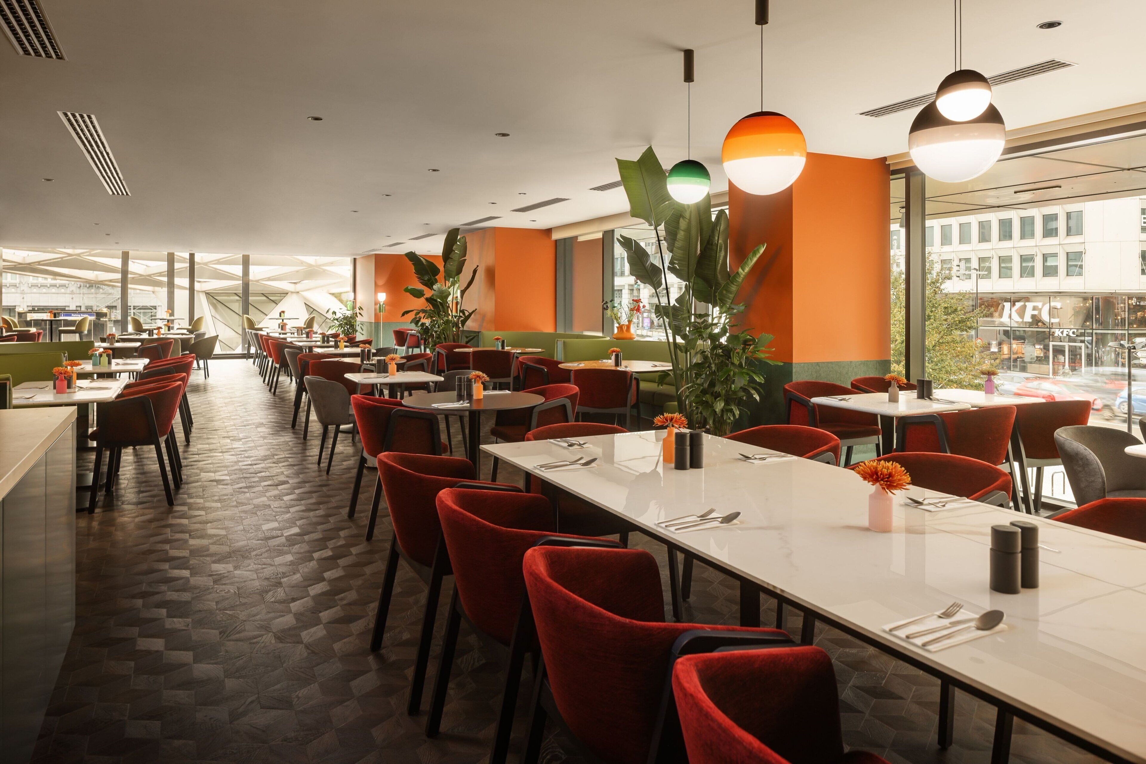 A modern restaurant with a long dining area, red chairs, and large windows offering a view of the city.