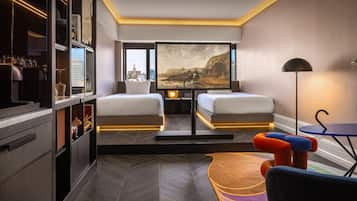 A modern hotel room with two beds, a large window with a city view, a desk with a lamp, and a colorful rug.