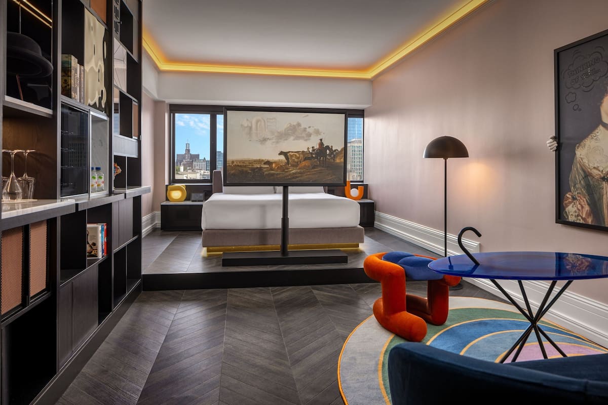 Ein modernes Hotelzimmer mit einem großen Bett, einem Schreibtisch, einem Bücherregal und Blick auf die Stadt.