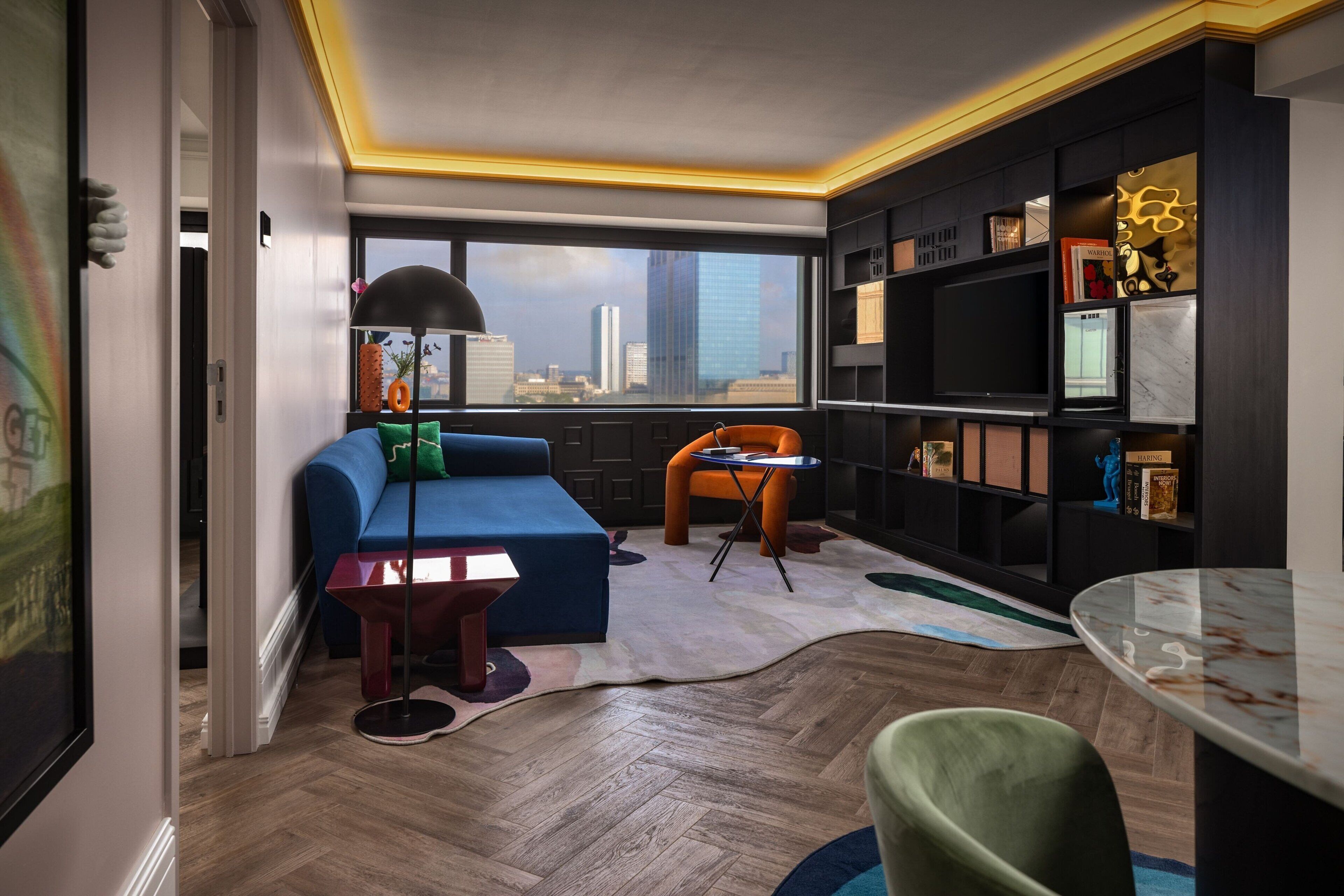 A modern living room with a blue sofa, orange chair, and a city view through large windows.