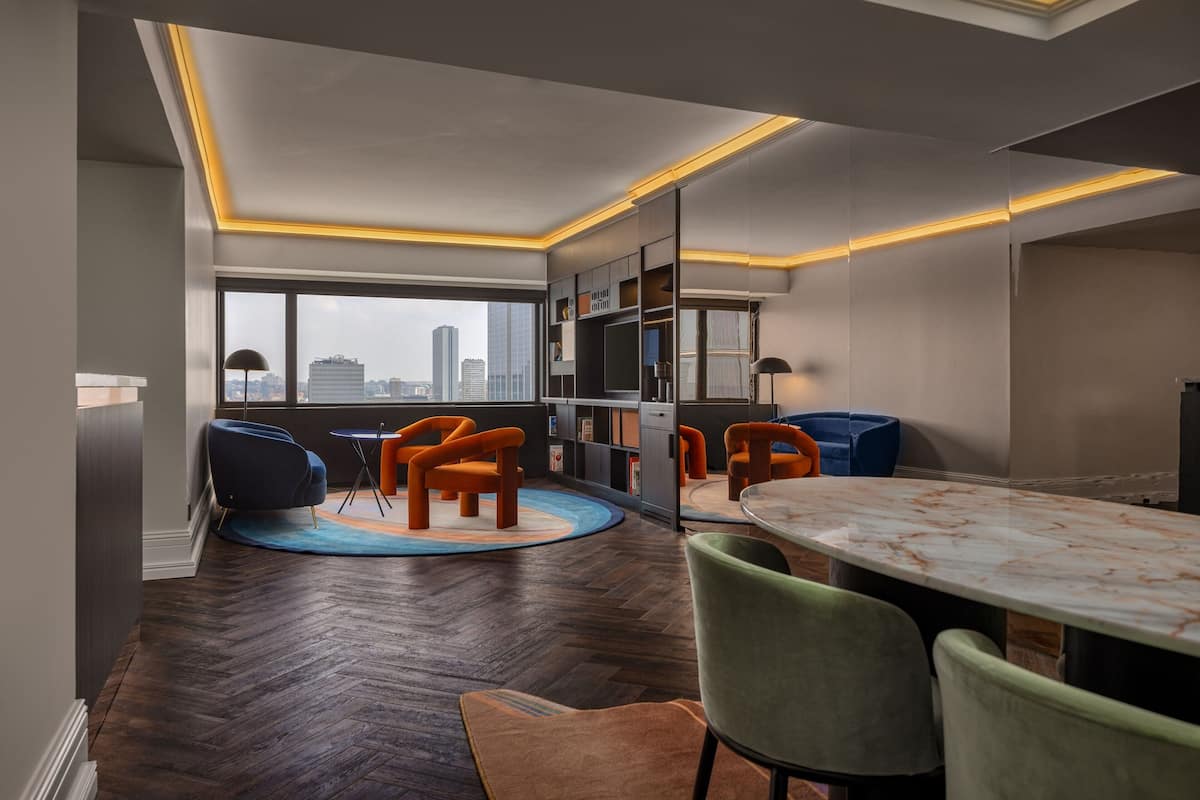 Ein modernes Hotelzimmer mit einem großen Fenster, einem runden Esstisch und orangefarbenen Stühlen.
