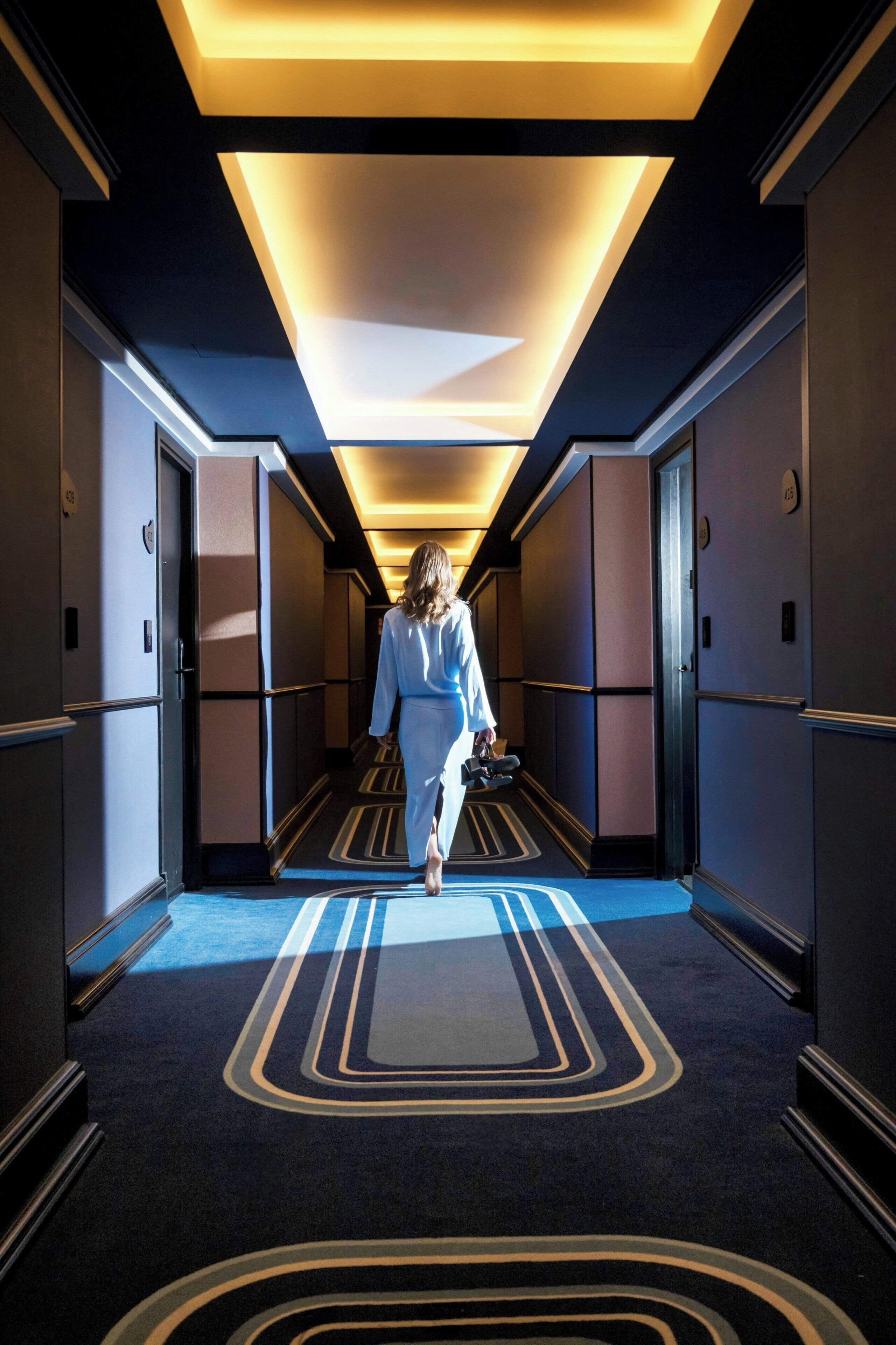 A modern hotel corridor with a geometric carpet design, a woman walking, and a contemporary ceiling light fixture.
