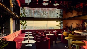 Un espacio lounge moderno con un sofá rojo, mesas redondas y taburetes amarillos, con amplias ventanas y vistas a la ciudad.