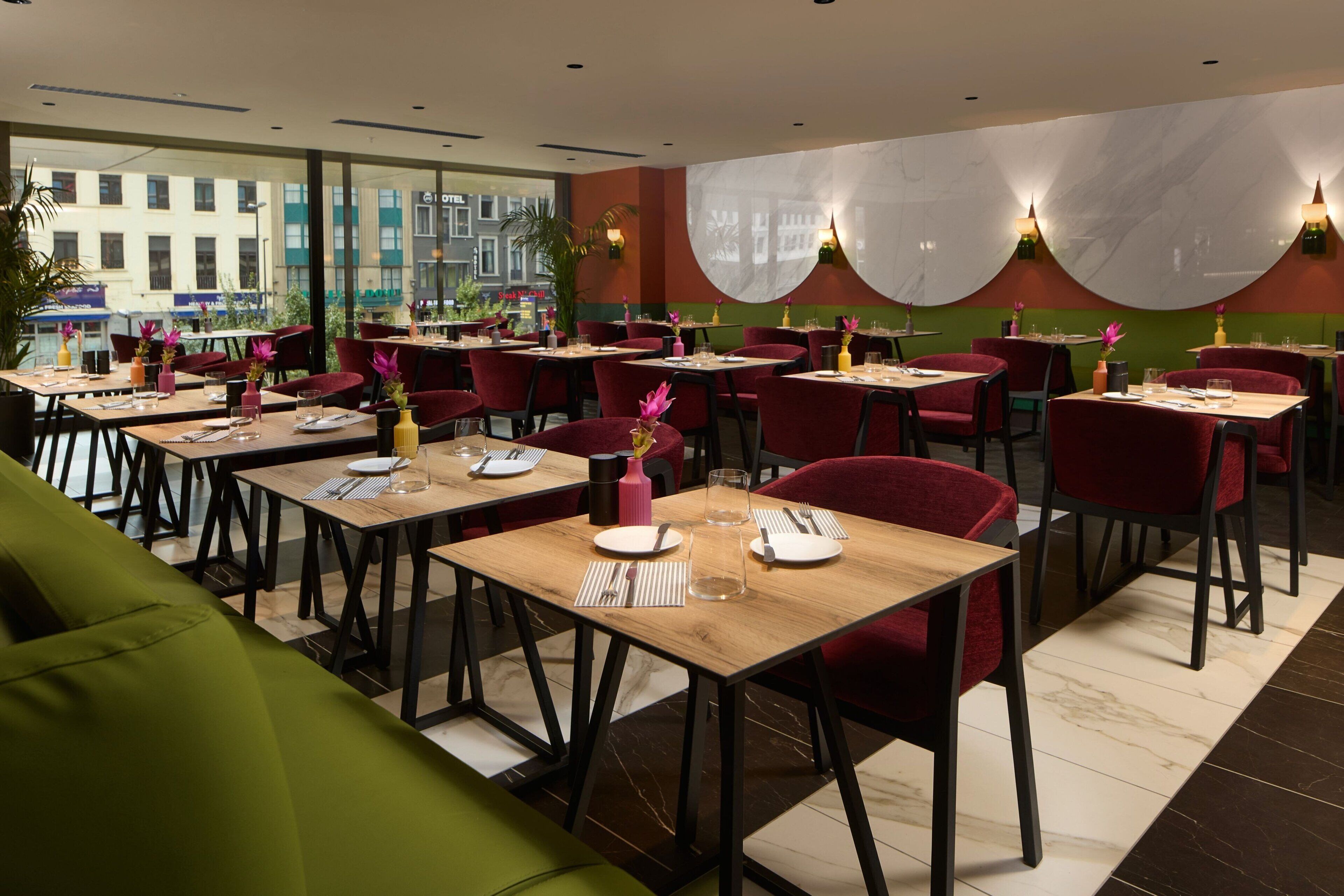 A restaurant with multiple tables, red chairs, and a green sofa. Tables are set with plates, cutlery, and glasses.
