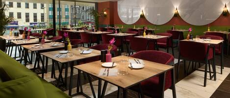 A restaurant with multiple tables, red chairs, and a green sofa. Tables are set with plates, cutlery, and glasses.