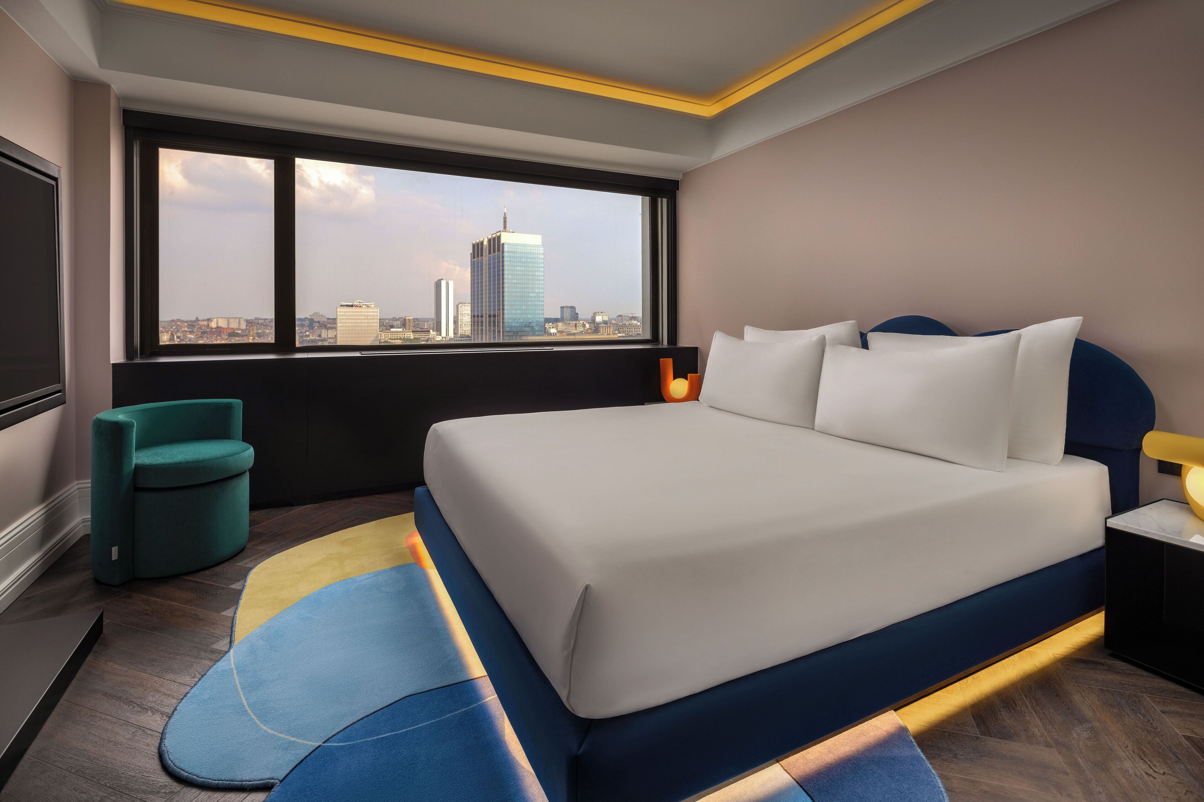 A modern hotel room with a large bed, a view of the cityscape, a teal armchair, and a small table with a lamp.