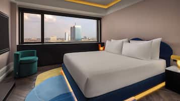 Ein modernes Hotelzimmer mit einem großen Bett, Blick auf die Stadtlandschaft, einem petrolfarbenen Sessel und einem kleinen Tisch mit einer Lampe.