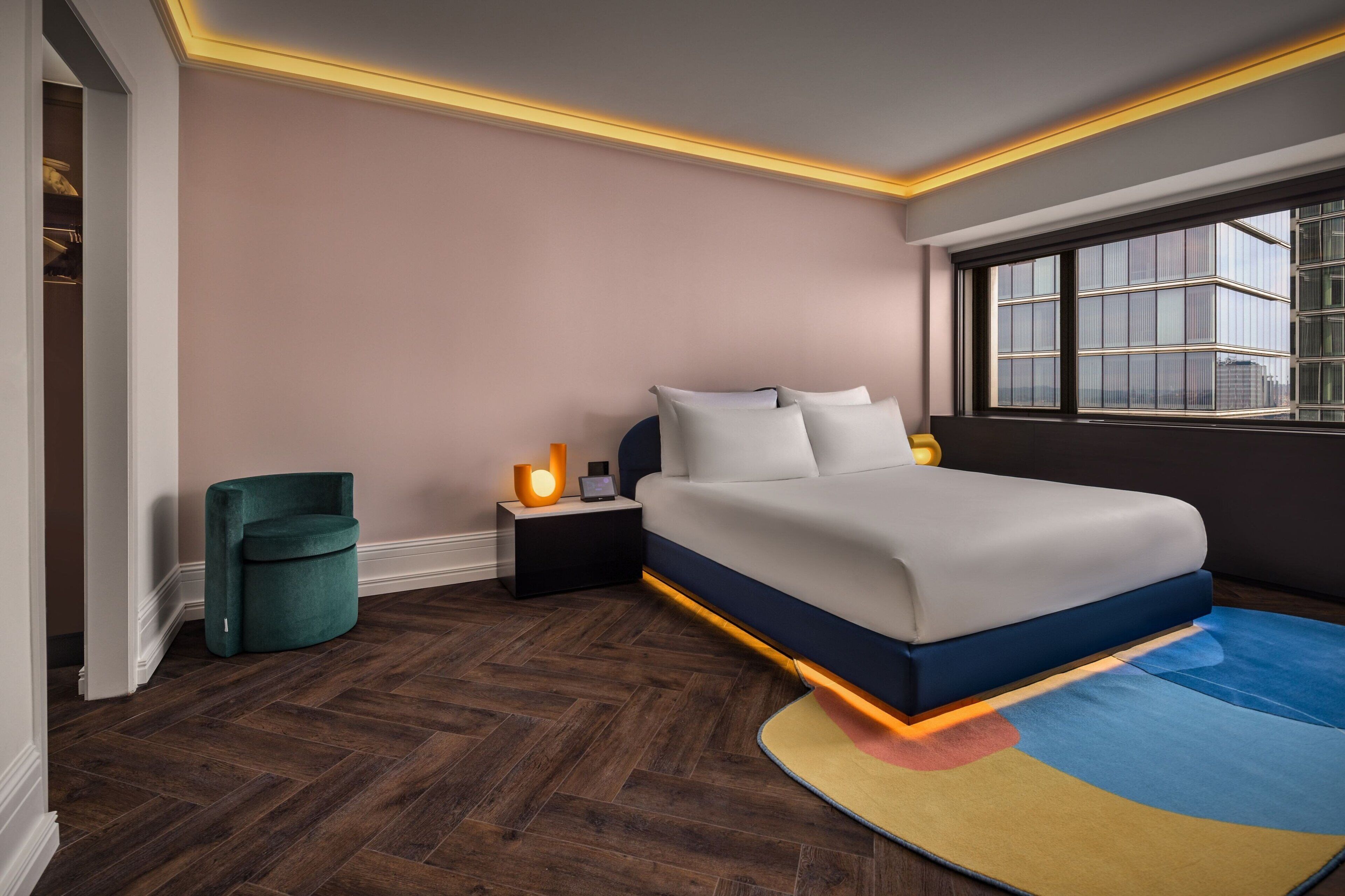 A modern bedroom with a large bed, a nightstand with a lamp, a green chair, and a window with a view of buildings.