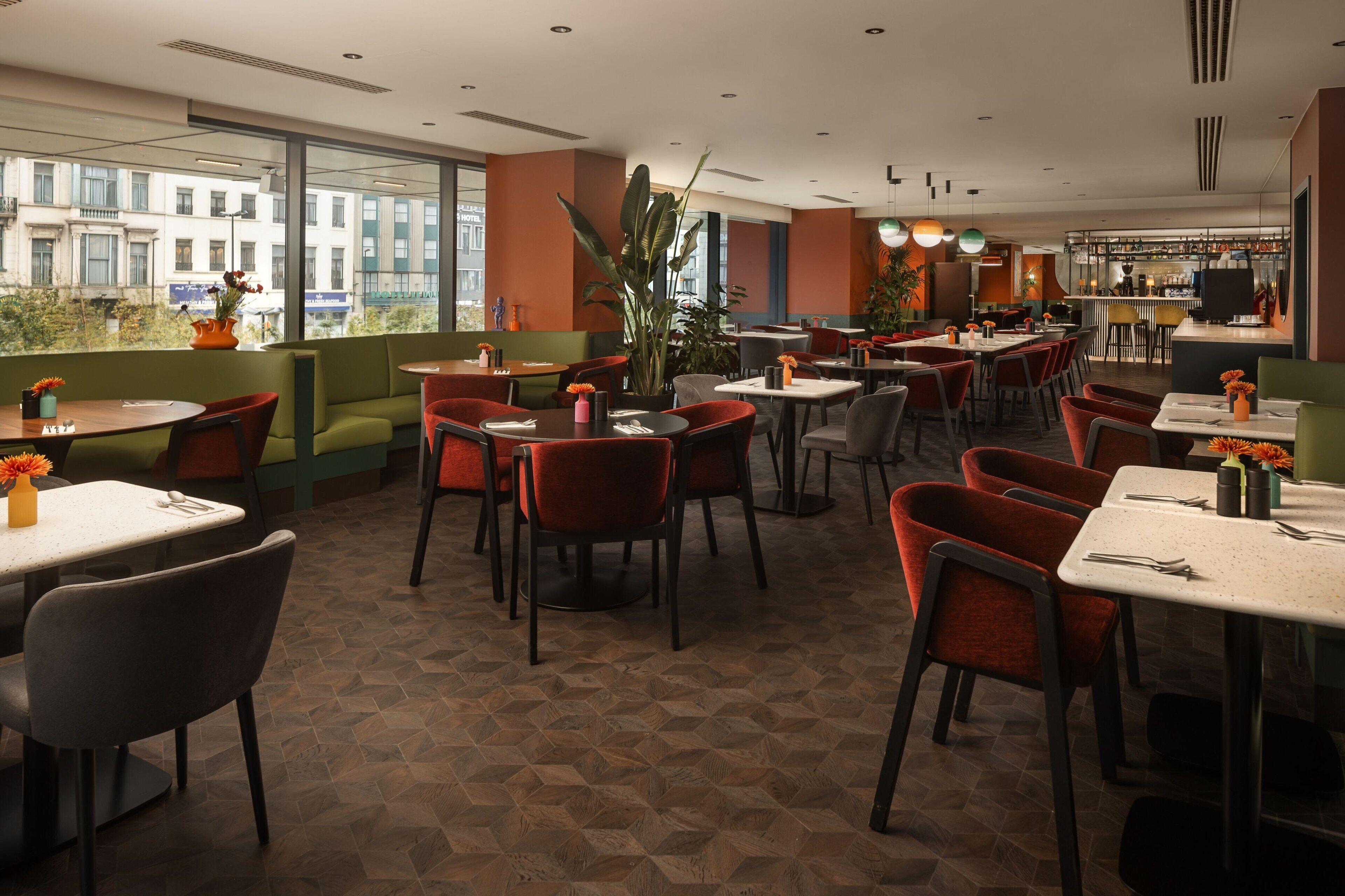 A restaurant with a mix of booths and tables, large windows offering a city view, and a bar area in the background.