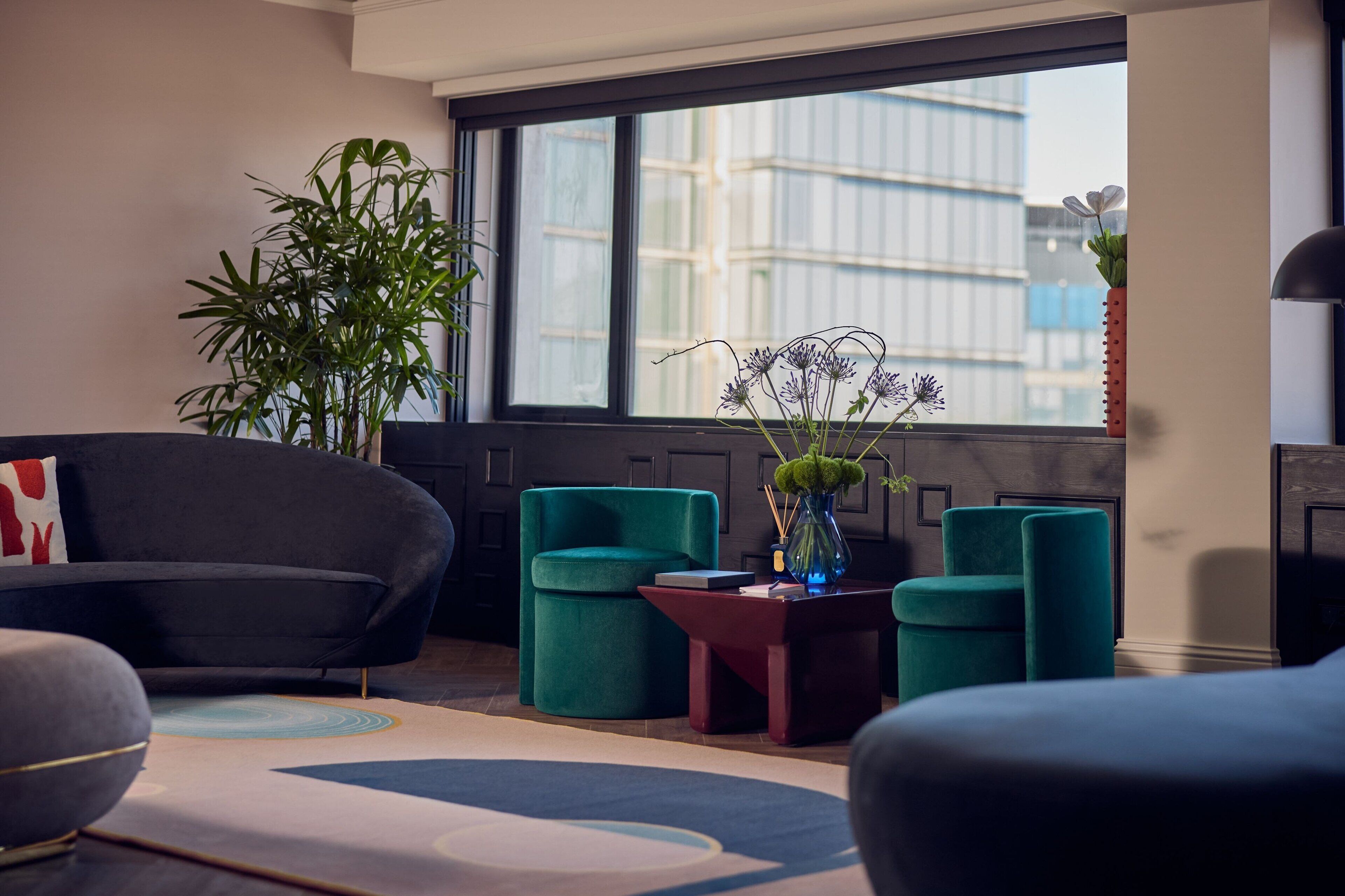 Un salón moderno con un ventanal grande, un sofá verde, sillones turquesa, una mesita de noche roja y un jarrón con flores.