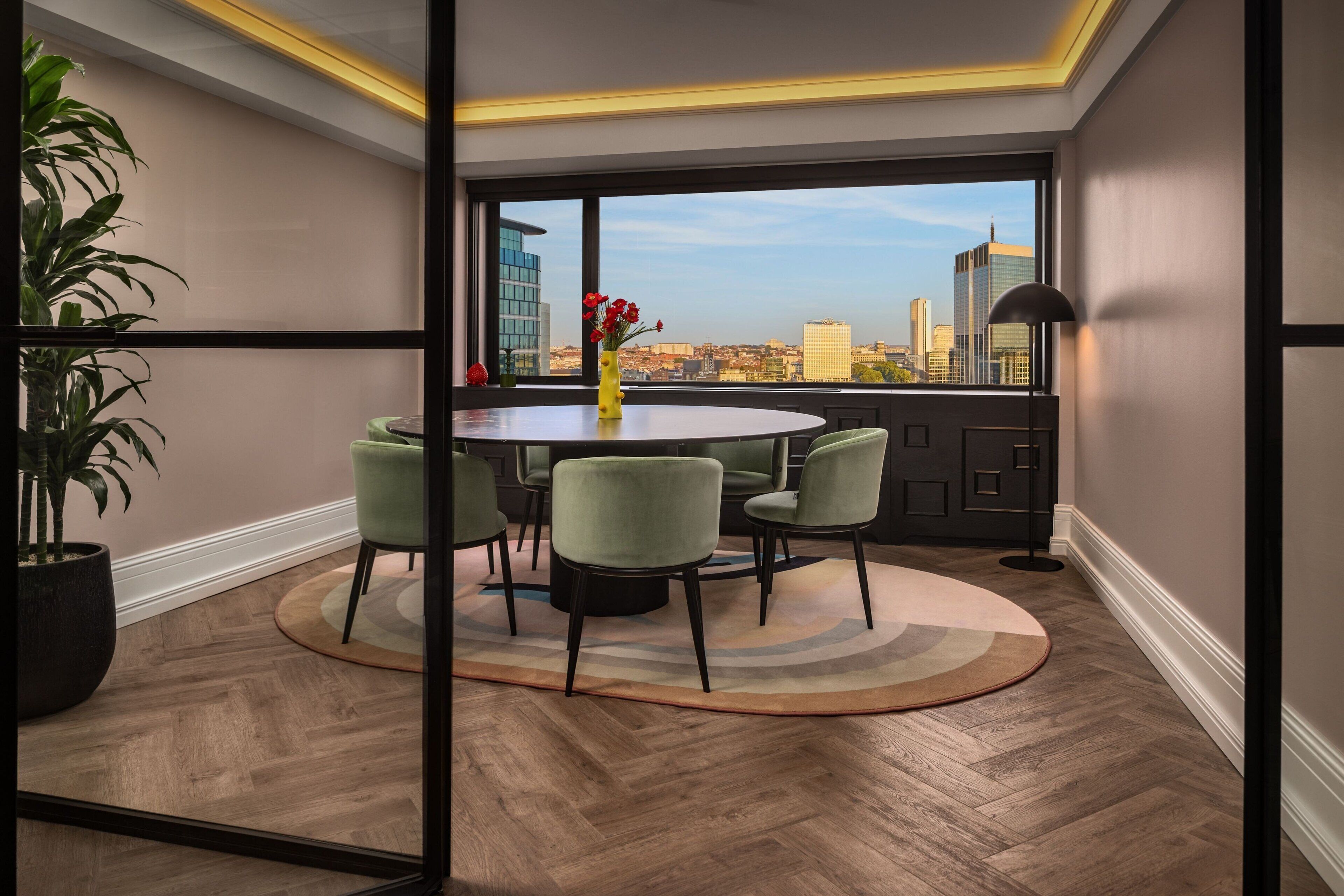 A modern dining area with a round table and six chairs, a potted plant, and a view of the city skyline through large windows.