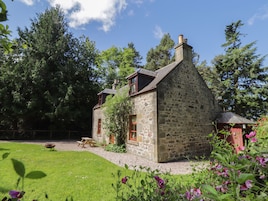 A stone house with a sloped roof, a chimney, and a small garden with flowers and a picnic table.