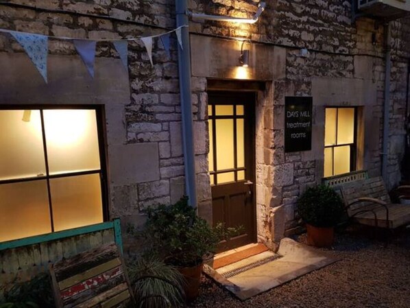A stone building with a sign indicating 'DAYS MIL treatment rooms'. There are two windows with lights on, a door, and a bench outside.