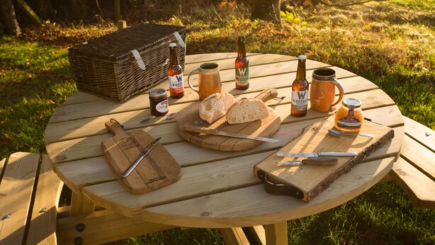 A picnic setup with a wicker basket, bottles, a mug, bread, and a knife on a wooden table. - The White Cottage (Elgin)