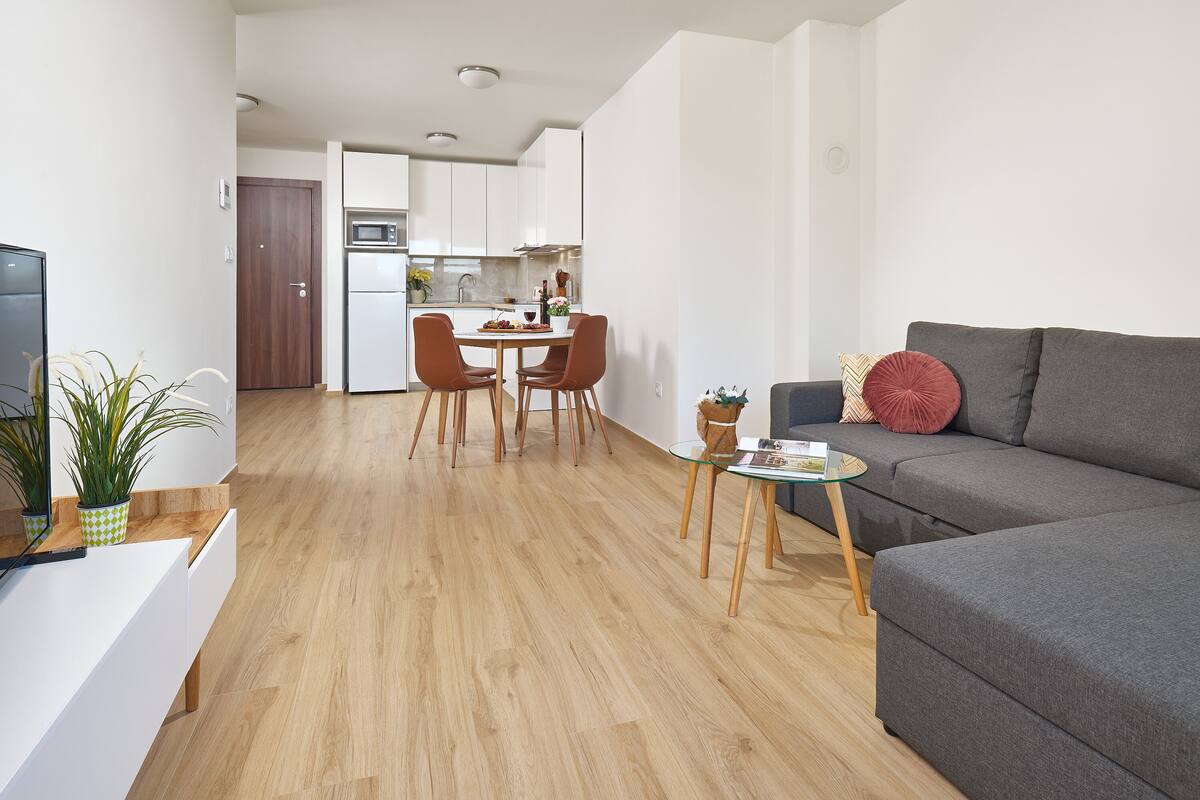 A modern living room with a grey sofa, a small glass coffee table, and a dining area with a white table and chairs.